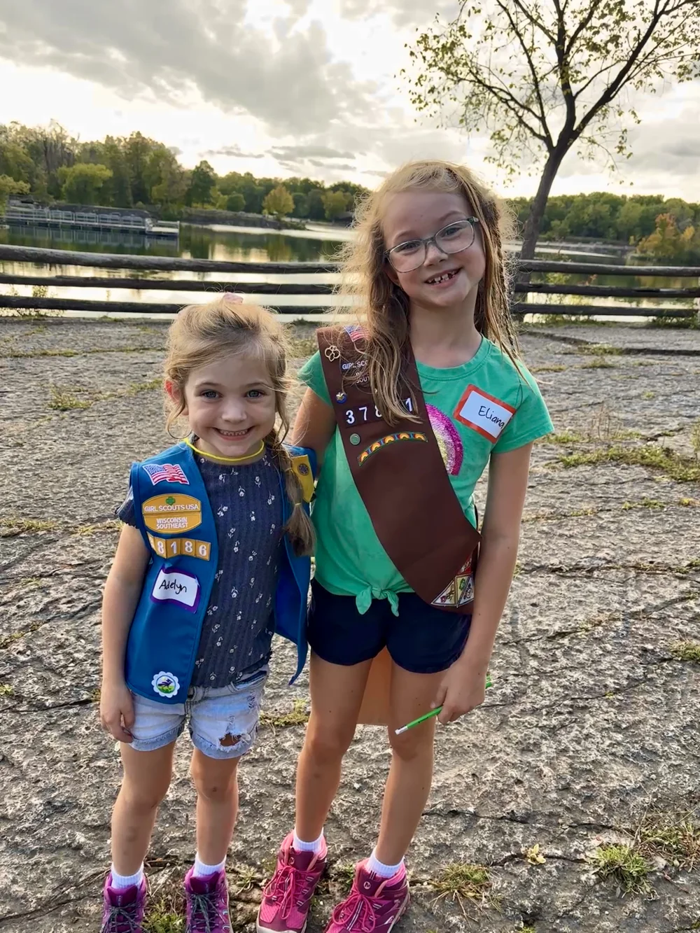 Addie in her blue Daisy vest and Ellie in her Brownie sash, smiling together outdoors